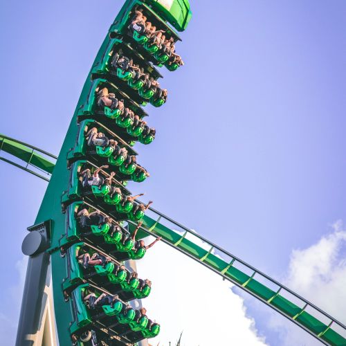 A green roller coaster is in steep descent with riders hanging upside down. The sky is clear with some clouds in the background.