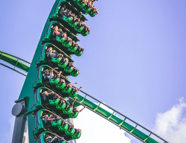 A green roller coaster is in steep descent with riders hanging upside down. The sky is clear with some clouds in the background.