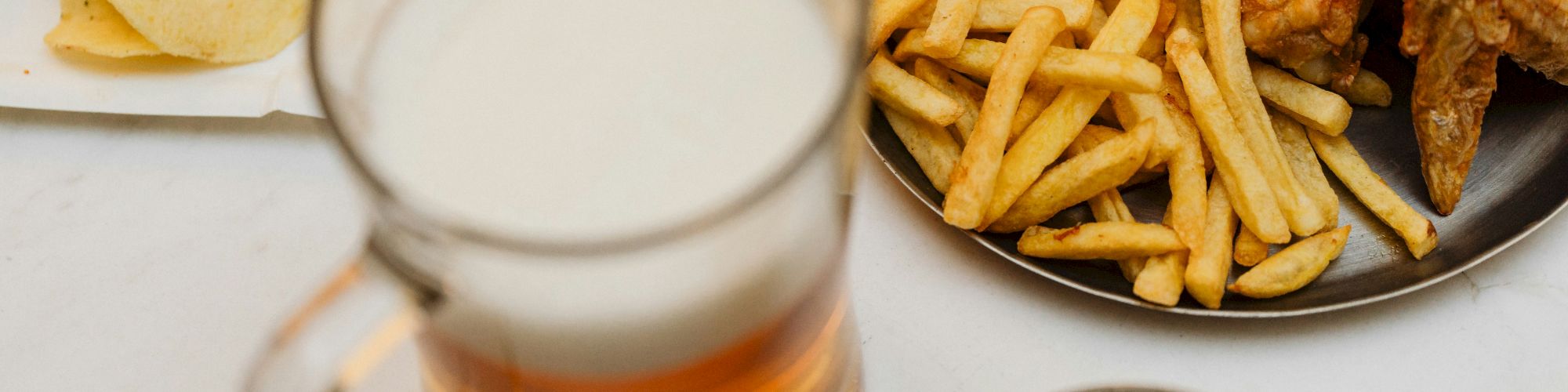 A table with a beer mug, fries and wings on a plate, a dish of pickles, and a small portion of chips with a burger.