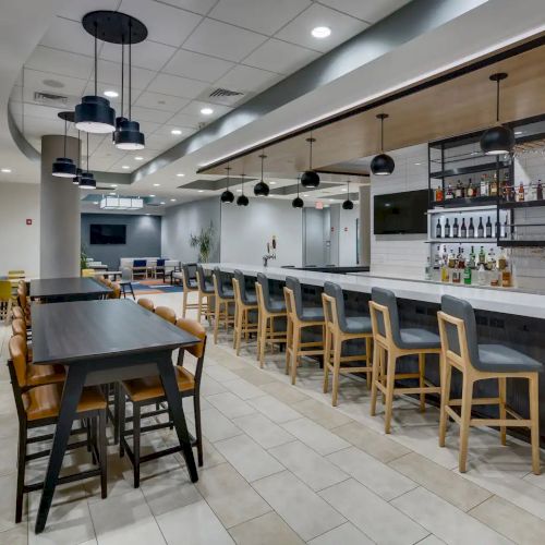 The image shows a modern bar with a long counter, high stools, a variety of drinks on shelves, and tables with chairs for dining.