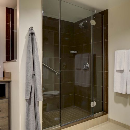 This image shows a modern bathroom with a glass shower, towels, a vanity with a mirror, and shelves with folded towels and a tissue box.