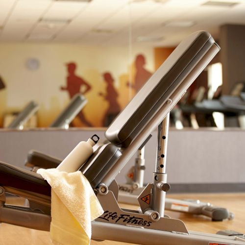 The image shows an adjustable gym bench with a towel and water bottle on it, set in a fitness room with mirrors and exercise equipment.