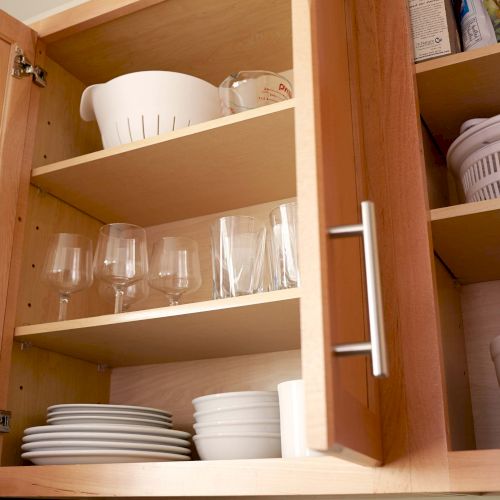 The image shows open wooden kitchen cabinets containing bowls, plates, glasses, a colander, and a salad spinner arranged neatly on shelves.