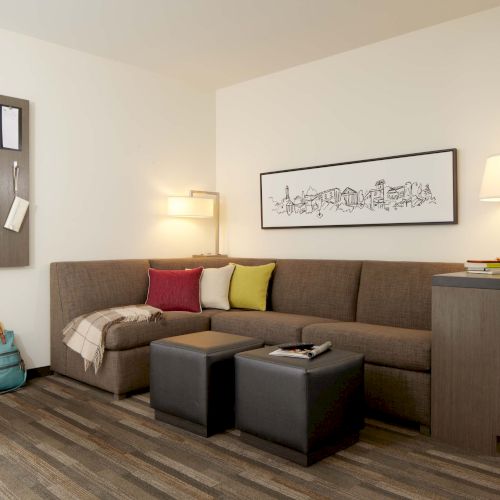 A cozy living room featuring a sectional sofa with colorful cushions, a lamp, two ottomans, a wall mirror, a framed picture, and a striped carpeted floor.