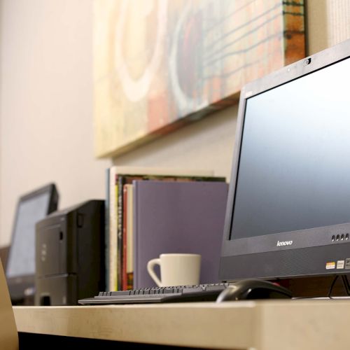 A computer workstation with a Lenovo monitor, keyboard, mouse, a coffee cup, a second workstation in the background, and art on the wall.