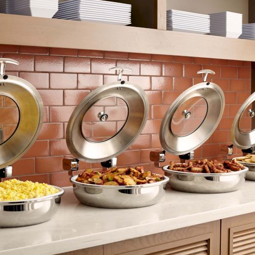 A buffet setup with dishes including scrambled eggs, roasted potatoes, sausages, and pancakes, against a brick backsplash.