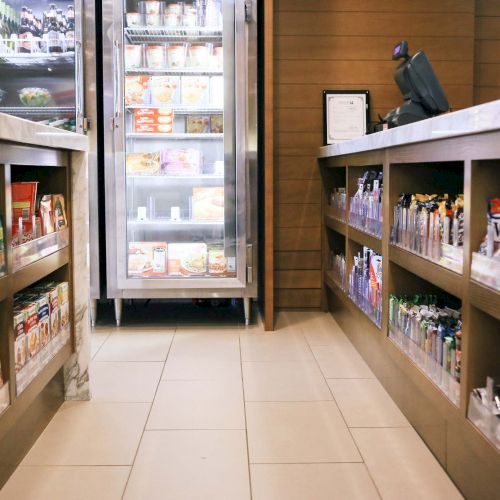 A small convenience store aisle with shelves stocked with various snacks and beverages; a refrigerated section is visible in the background.