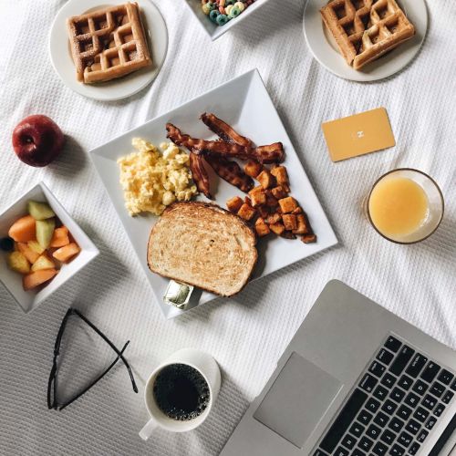 An appetizing breakfast spread with waffles, fruits, scrambled eggs, toast, a laptop, coffee, juice, and eyeglasses on a white surface.