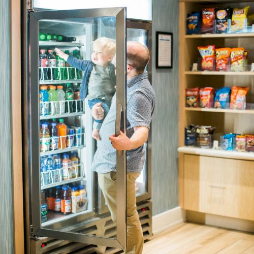 A man is holding a child who is partially inside a drink refrigerator in a store. Shelves with snack items are visible on the right side of the image.