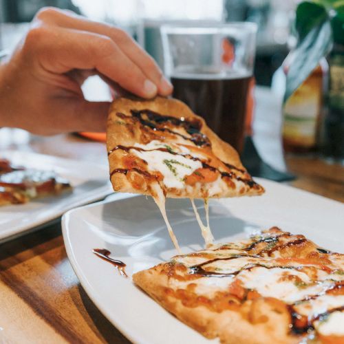 A person is lifting a slice of pizza from a plate. Another plate and a glass of drink are also visible on the table in the background.