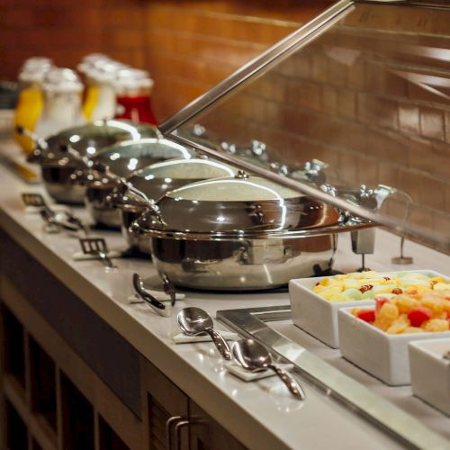 A buffet setup with chafing dishes, trays of fruit, and various beverages in dispensers is on a counter with utensils and serving spoons.