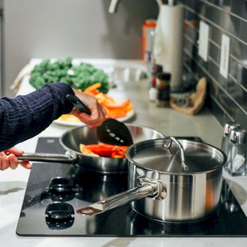 A person is cooking in a kitchen using two stove top pans, with various ingredients and utensils nearby.