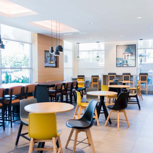 A modern, well-lit cafe interior with round tables and mix-matched chairs, featuring pendant lights and large windows.