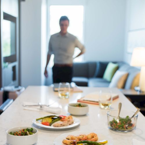 A kitchen with plates of food and bowls of salad on a countertop, with a person standing in the background near a sofa.