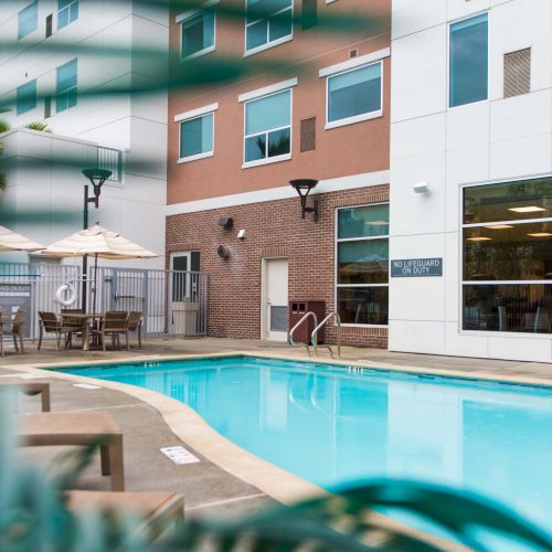 An outdoor pool with lounge chairs, umbrellas, and a building with large windows and a sign in the background.