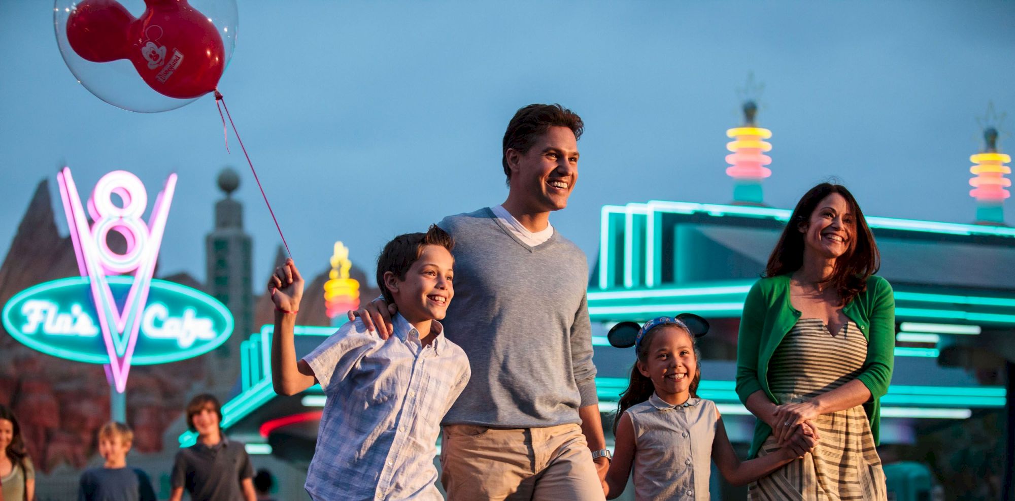 A family walks happily at an amusement park, with neon lights and balloons creating a festive atmosphere.