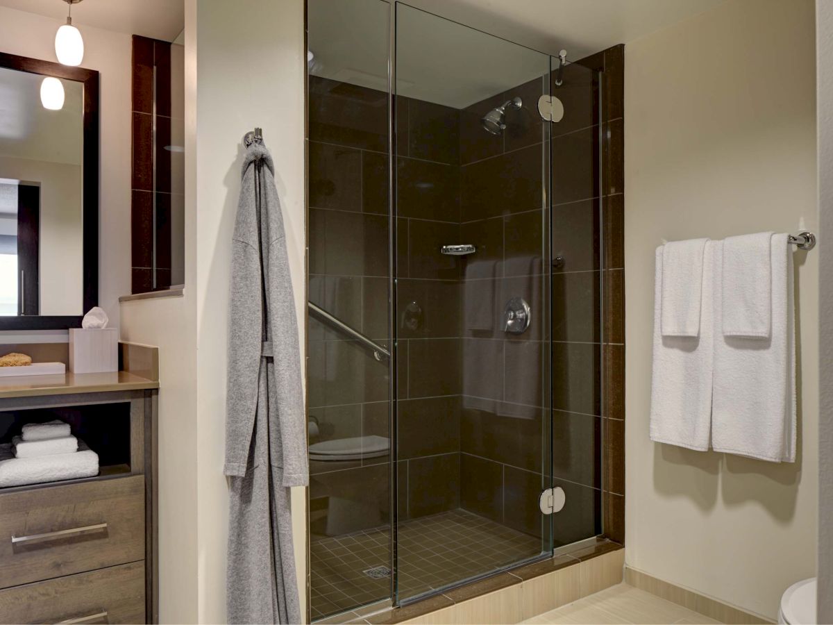 This image shows a modern bathroom with a glass-enclosed shower, towels hanging on a rack, a robe on a hook, and a wooden vanity with a mirror.