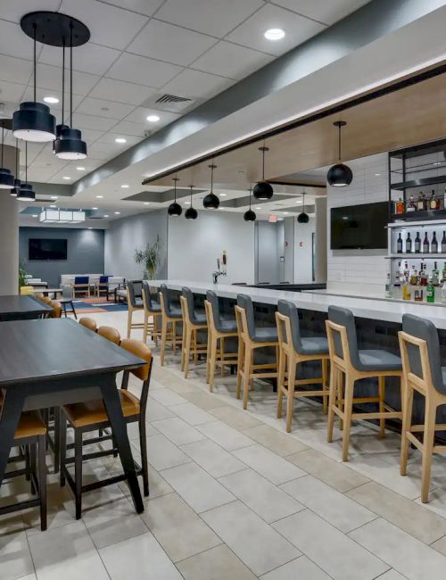 A modern bar and lounge area with high tables and bar stools, pendant lighting, and a fully stocked bar in a spacious, well-lit room.