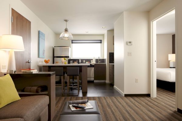 A modern apartment with an open kitchen, dining area, and living space. A bedroom is partially visible through a door on the right.