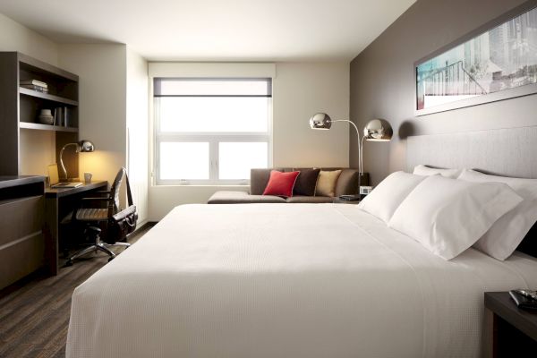 A modern hotel room with a neatly made bed, desk with chair and lamp, sofa with pillows, window with blinds, and a wall art above the bed.