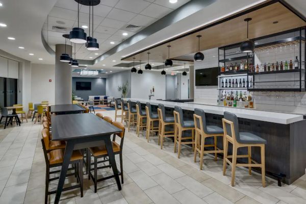 The image depicts a modern bar area with a long counter, bar stools, tables, and chairs. Bottles and glasses are displayed behind the counter.