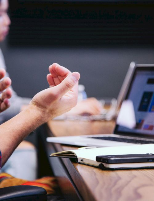 A person gestures while discussing in front of a laptop in a meeting room, with a notebook and phone on the table in front of them.