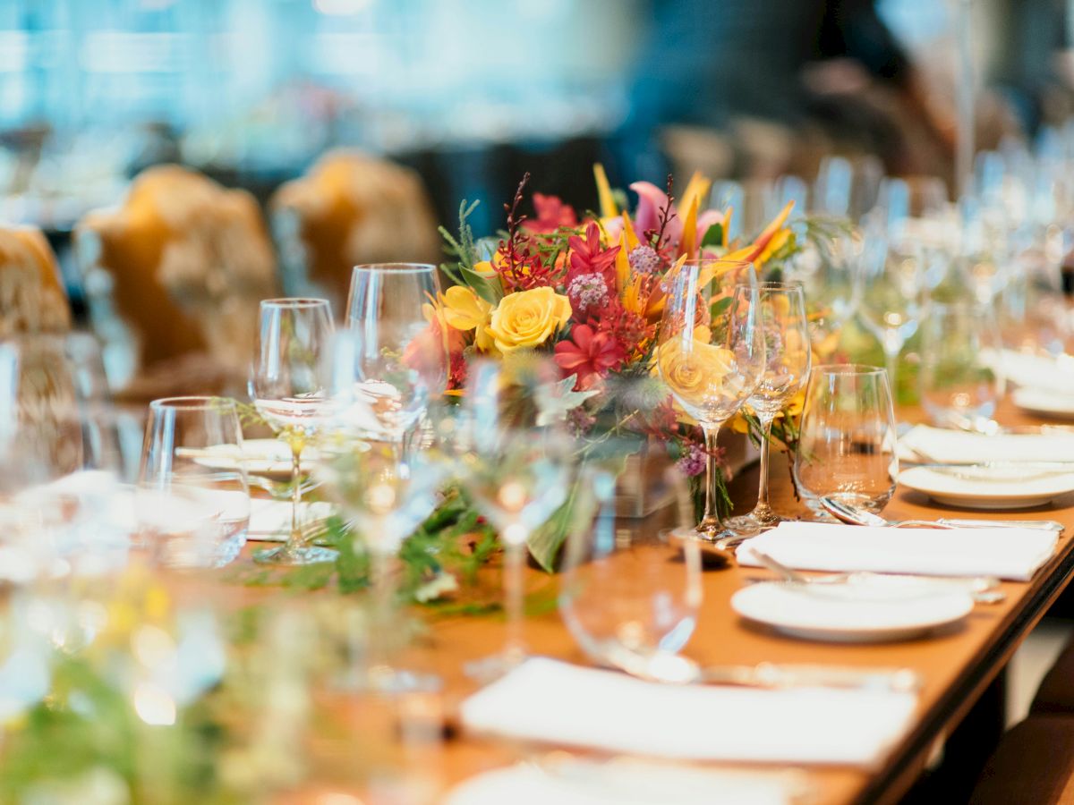 A long dining table is elegantly set with white plates, clear glasses, and colorful flower centerpieces, ready for a formal event or celebration.