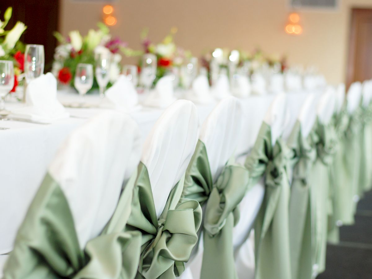 The image shows a table set up for a formal event, with white chair covers and green bows, flowers, glasses, and napkins arranged neatly.