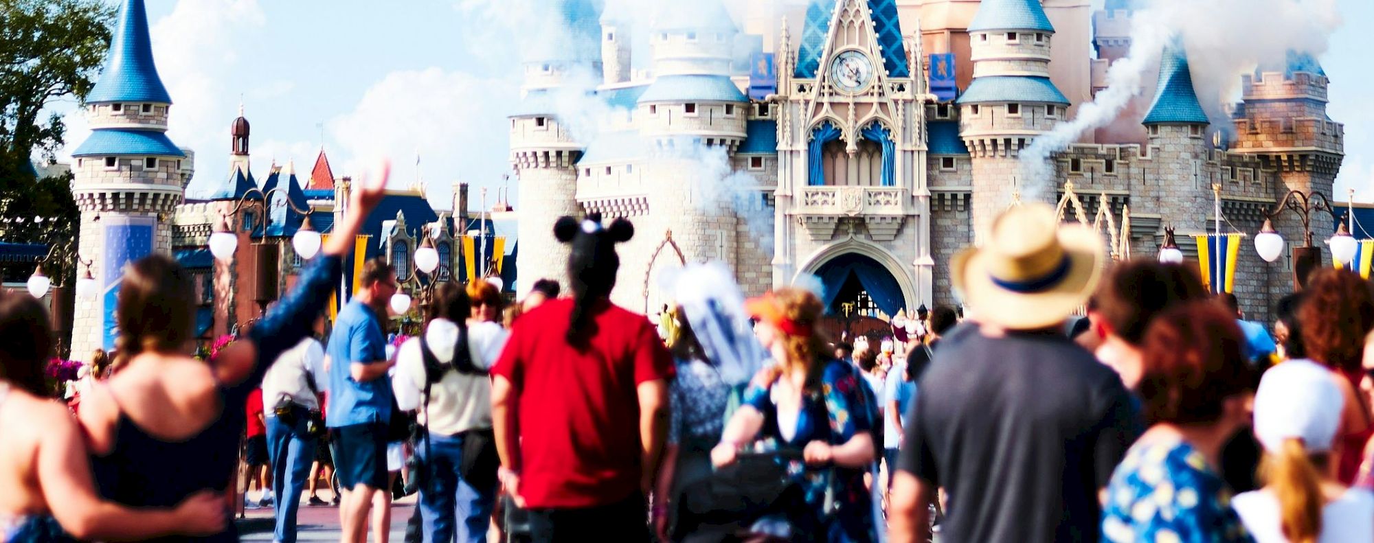 A crowd of people gathers in front of a majestic castle with fireworks streaming across a clear blue sky in what appears to be a theme park.