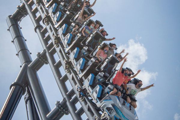 The image shows a group of people riding a steep, thrilling roller coaster against a blue sky with some clouds, hands up in excitement.