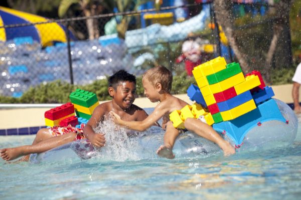 Two kids are playing in a pool on a float made of large, colorful LEGO pieces, splashing water and smiling happily.