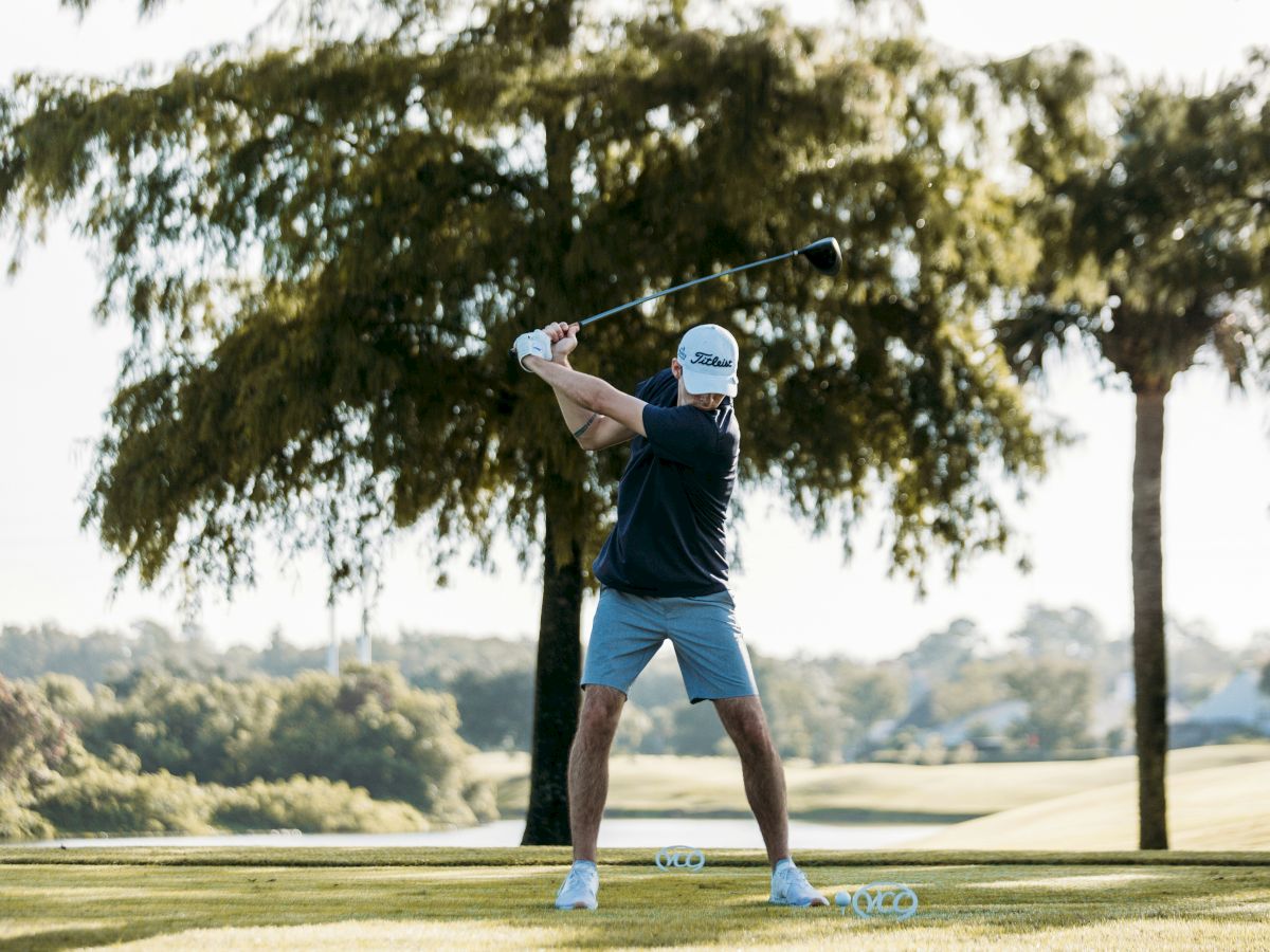 A person wearing a cap and shorts is taking a golf swing on a lush, green golf course, with trees and a clear sky in the background.