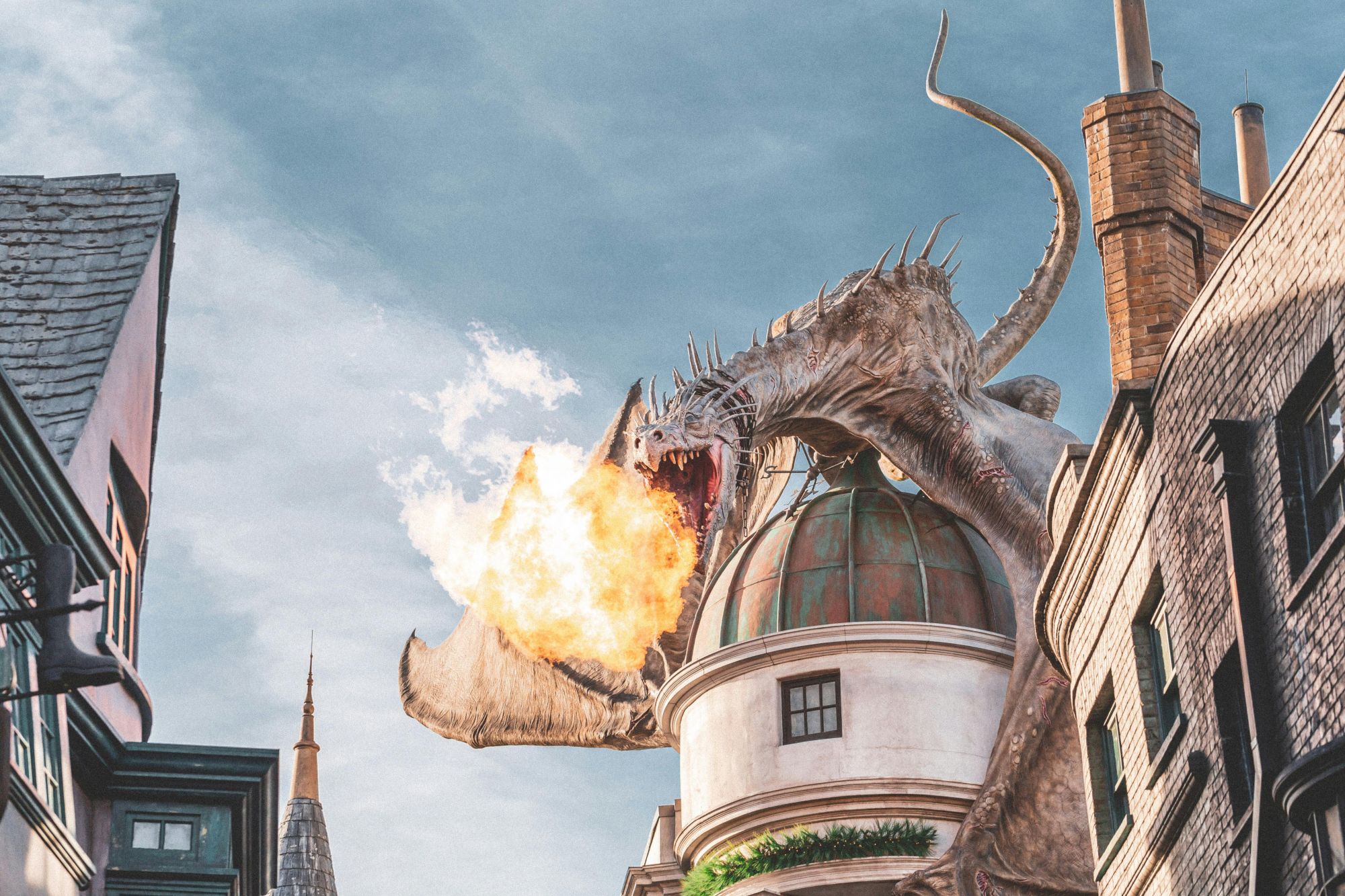 A dragon perched on a rooftop is breathing fire amidst fantasy-style buildings under a clear sky.